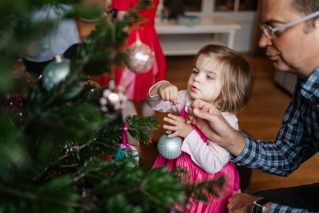 A little girl trying to hand a ornament on the Christmas tree with her daddy's helpp